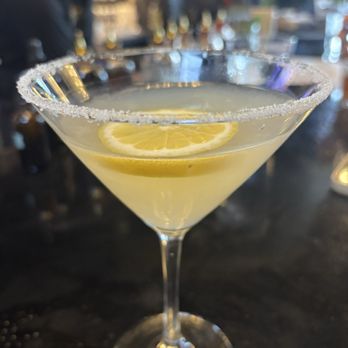 Bistro 135 - A lemon martini with a salted rim and lemon slice garnish is placed on a bar counter, with a blurred bar scene in the background. - Italian Near Me - Tracy, California