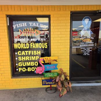 Fish Tales - The image shows the storefront of Fish Tales in Corrigan, TX, with a yellow brick exterior. Two large windows display the business name, hours, and a fish-themed logo. A colorful flamingo decoration and a multi-colored welcome sign are placed outside near a potted plant, creating a welcoming entrance. The business hours are listed on the door, indicating operating times from Monday to Sunday. - Comfort Food Near Me - Corrigan, Texas