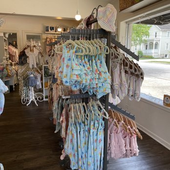 A Baby Naturally - Children's clothing such as dresses, rompers, and outfits are displayed on racks inside a boutique store. The store has large windows allowing natural light, with a view of residential houses outside. The interior features wooden flooring and a variety of clothing items arranged neatly on hangers, with some racks positioned near the window and others further inside. The store appears organized and dedicated to natural or organic children's apparel. - Baby Gear & Furniture Near Me - Wheaton, Illinois