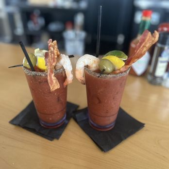 The Point - Bloody Mary's $14 with bacon, shrimp and Tito's. - Two Bloody Mary drinks with shrimp, bacon, olives, and lemon garnishes sit on black napkins at a bar. - American Near Me - Marshfield, Massachusetts