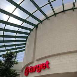 The image shows the exterior of a Target store, featuring a curved beige tiled wall with a large, illuminated red Target logo. Above, there is a glass ceiling with green framing, allowing natural light to enter the space. A small tree is visible in the lower left corner, adding greenery to the scene.