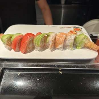 Koi Roll - Sushi combination - A sushi roll featuring different toppings such as avocado, salmon, shrimp tempura, and possibly crab, arranged in a line on a white rectangular plate. The plate is placed on a metal surface, with a person in black clothing visible in the background. - Sushi Bars Near Me - Rolla, Missouri
