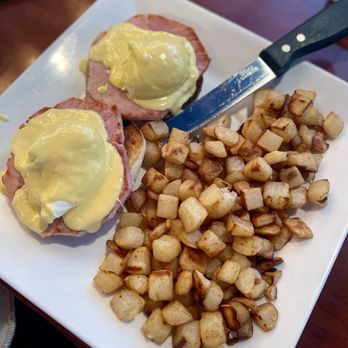 Shine Cafe - Eggs Benedict - A white square plate holds two Eggs Benedict with ham and hollandaise sauce, accompanied by a large portion of diced, browned potatoes. A knife rests on the plate, and the background shows a wooden table surface. The dish is served in a casual brunch setting at Shine Cafe in Troy, Michigan. - Breakfast & Brunch Near Me - Troy, Michigan