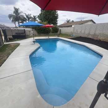Cerda Pool Construction - A backyard swimming pool with a concrete deck is shown, featuring umbrellas in red, blue, and red providing shade. The pool is surrounded by a fence, with neighboring houses and palm trees visible in the background. The sky is partly cloudy, and the area appears to be a private residential yard. - Pool & Hot Tub Service Near Me