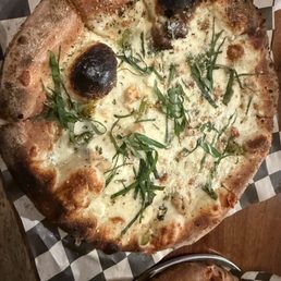 White pie - A pizza featuring a white cheese base, garnished with fresh green herbs, and a crust with some charred areas. The pizza is placed on black and white checkered paper, likely in a restaurant setting.