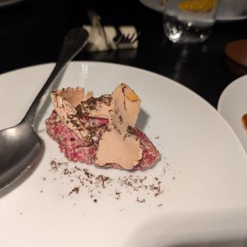 Asador Bastian - Tartare - A white plate holds a serving of beef tartare topped with slices of foie gras and sprinkled with black truffle shavings. A metal spoon rests on the plate's edge. The background includes a glass of water, a brown leather item, and other plates, indicating a restaurant environment. The presentation emphasizes fine dining and gourmet ingredients. - Spanish Near Me - Chicago, Illinois