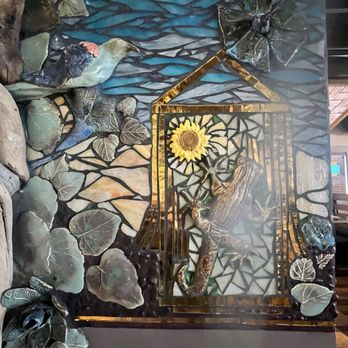 Range Cafe - The image shows a mosaic artwork composed of various materials including metal, ceramic, and possibly glass. It features a frog positioned near a sunflower, with surrounding leaves and insects, all set against a dark background with a textured surface. The mosaic includes detailed elements like a butterfly and a dragonfly, with the frog appearing to be climbing or resting among the foliage. The artwork is framed with a metallic border and is placed on a flat surface, possibly in an indoor setting. - Breakfast & Brunch Near Me - Albuquerque, New Mexico