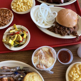 Bartley's Bar-B-Que - Pulled Pork, Mac and cheese - A meal tray features smoked barbecue meats, including ribs and shredded beef, accompanied by sides such as macaroni and cheese, baked beans, and sliced onions. Additional sides include pickled vegetables and a bun with shredded beef. The tray is set on a wooden table, with a small cup of sauce and a wrapped dessert also visible. The presentation suggests a casual barbecue restaurant setting. - Barbeque Near Me - Grapevine, Texas