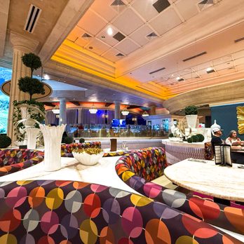 Biscotti's - View toward a bar area - The image shows the interior of a restaurant with a curved ceiling and warm lighting. Colorful, patterned upholstered seating surrounds marble tables, with decorative vases and topiary plants on display. In the background, a bar area with stools and a blue-lit display is visible, along with a few diners seated at tables. The decor includes large mirrors and ornate accents, creating an upscale atmosphere. - Cafes Near Me - Reno, Nevada