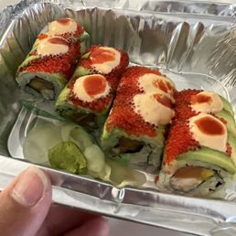 Inferno roll - Four sushi rolls are presented in a foil container, each topped with fish roe, mayonnaise, and a spicy orange sauce. The rolls contain visible ingredients such as avocado and possibly fish or seafood. Sliced cucumber and a dollop of wasabi are placed beside the sushi. The presentation suggests a takeout or delivery order from a sushi restaurant.