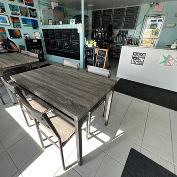 Kool Beenz Coffee - The image shows the interior of a coffee shop with two wooden tables and metal chairs arranged for customers. The shop has a chalkboard menu on the wall behind the counter, which displays various drink options. The counter area includes coffee-making equipment and a small chalkboard sign. Sunlight enters through a door with a window, casting shadows on the tiled floor. The decor includes artwork on the walls and a black mat near the entrance. - Coffee & Tea Near Me - Flagler Beach, Florida