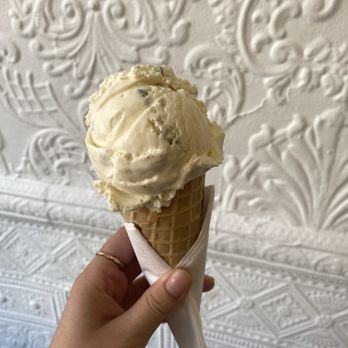 Malai - Peda - A person holds a vanilla ice cream cone with a waffle cone and a napkin, with a textured white decorative wall in the background. - Ice Cream & Frozen Yogurt Near Me - Brooklyn, New York