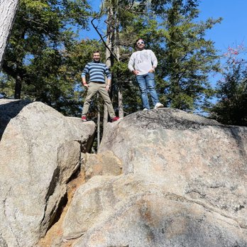 Lower Falls - Two men are positioned on large, uneven rocks in a forested area, surrounded by tall trees with green foliage. One man is wearing a striped sweater and khaki pants, while the other is dressed in a light-colored sweatshirt and jeans. Both are wearing sunglasses and appear to be enjoying an outdoor activity on a sunny day. - Parks Near Me - Conway, New Hampshire