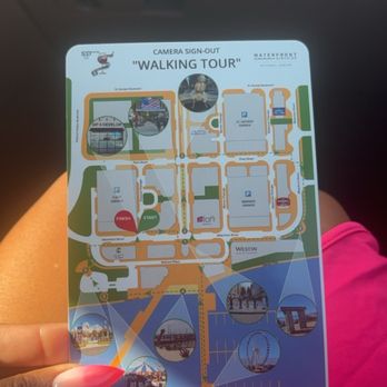 Sip & Develop - A person with pink-painted nails is holding a map titled 'Walking Tour' inside a vehicle. The map features a layout of a location with various points of interest, pathways, and parking areas. The background shows part of the vehicle's interior, and the person is wearing a pink garment. The map includes images of landmarks and a route for a walking tour. - Event Photography Near Me - Oxon Hill, Maryland