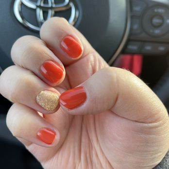 High Tech Nails - Thanksgiving gel nails - A close-up of a person's hand displays freshly painted nails, with orange polish on most fingers and a single accent nail covered in gold glitter. The hand is positioned in front of a Toyota steering wheel, indicating the photo was taken inside a car. The background includes part of the vehicle's dashboard and controls, with natural lighting illuminating the nails. - Nail Salons Near Me - Normal, Illinois