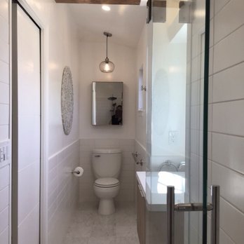 Method Remodeling - Toilet, vanity and medicine cabinet. - The bathroom has a narrow layout with white tiled walls on the lower half and smooth white paint on the upper half. A toilet is positioned at the back, with a small vanity and sink to the right. A mirror is mounted above the toilet, and a medicine cabinet is visible on the wall. The ceiling includes a wooden beam and recessed lighting, with a pendant light hanging from the ceiling. A glass shower enclosure is on the right side, with metal hinges and handles. The floor is covered with light-colored marble or tile. The space appears clean and modern, with a minimalist design. - General Contractors Near Me - Burlingame, California