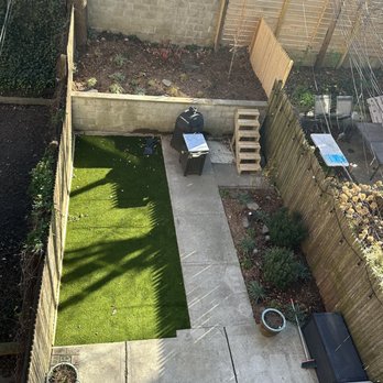 Native NY Gardens - After - A backyard features a concrete patio area with a small patch of artificial grass on one side. Garden beds with plants are enclosed by wooden fences and a brick wall. Gardening supplies, including a barbecue grill, a small table, and potted plants, are visible. The space is divided into sections with fencing and a wooden pallet, with sunlight casting shadows across the yard. - Landscaping Near Me