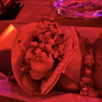 Cave Restaurant - Tacos - Two tacos with toppings are served on a white plate in a dimly lit restaurant with red lighting, alongside a glass of water. - Lounges Near Me - Brooklyn, New York