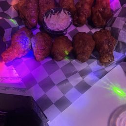 Spicy Chicken Wings - A serving of fried chicken wings is displayed on checkered paper, with a small bowl of dipping sauce placed in the middle. The wings are arranged in two rows, with five wings in each row. The lighting in the setting is purple and green, creating a colorful ambiance. The image appears to be taken in a casual dining or bar environment.
