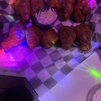 Echo Box - Spicy Chicken Wings - A serving of fried chicken wings is displayed on checkered paper, with a small bowl of dipping sauce placed in the middle. The wings are arranged in two rows, with five wings in each row. The lighting in the setting is purple and green, creating a colorful ambiance. The image appears to be taken in a casual dining or bar environment. - Karaoke Near Me - Washington, DC, District of Columbia