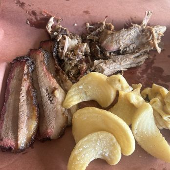 Sacred Ground - Brisket, pulled pork, mac and cheese, and fries - The image shows slices of smoked brisket, shredded pulled pork, and crispy fried potato chips arranged on a brown paper-lined surface. The brisket has a dark, caramelized crust, while the pulled pork appears tender and moist. The potato chips are golden and salted. The background includes a wooden table and a small candle or container in the upper right corner. - Salad Near Me - Flora, Mississippi