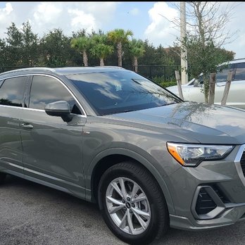 Jonathan Window Tint - A gray Audi SUV is parked in a parking lot, with trees and other vehicles visible behind it, under a partly cloudy sky. - Home Window Tinting Near Me - Boca Raton, Florida