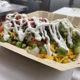 Veg Chorizo Burrito Bowl - A takeout container holds a beef burrito bowl topped with sour cream, shredded cheese, chopped green onions, pico de gallo, guacamole, and roasted red peppers. The bowl contains rice, beans, and other ingredients, with a drizzle of sour cream on top. The background shows a kitchen setting with stainless steel surfaces and appliances.