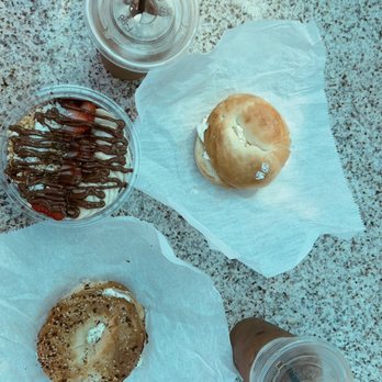 Beaches & Cream - Açaí bowl, regular and everything bagels with Cape Cod  chip cream cheese, and iced coffees! - The image shows two bagels placed on white parchment paper on a granite table, one topped with seeds and the other plain. There is a small container of chocolate-covered strawberries and two iced drinks in clear plastic cups with black straws. The table surface is speckled gray and brown, and the drinks appear to be iced coffee or tea. - Juice Bars & Smoothies Near Me - West Dennis, Massachusetts