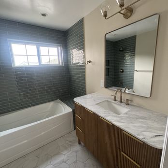 Method Remodeling - The bathroom includes a white bathtub positioned beneath a window with multiple panes, allowing natural light. Adjacent to the bathtub is a shower area with gray subway tiles. The vanity has a marble countertop with an integrated sink, a modern faucet, and a wooden cabinet with vertical grooves and gold handles. Above the vanity is a large, slightly tilted mirror, and a three-bulb light fixture with glass shades provides additional lighting. - General Contractors Near Me - Burlingame, California