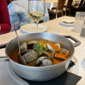 Sagres Restaurant - Littlenecks - A metal pot filled with steamed clams in broth is placed on a table at a restaurant, accompanied by a glass of white wine and dining utensils. - Portuguese Near Me - Fall River, Massachusetts