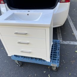 Bathroom vanity - A white bathroom vanity with a built-in sink and three drawers is positioned on a blue plastic rolling cart in a parking lot.