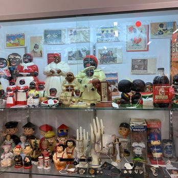 Antiques On Pierce - racist and offensive art and figures - The image shows a glass display case filled with vintage and collectible items such as figurines, dolls, sculptures, and memorabilia. The top shelf features numerous black and white and colorful figurines, some dressed in traditional or historical attire, along with photographs and posters in the background. The lower shelf contains more figurines, including some in traditional clothing, as well as jewelry and small objects. The display appears to be part of an antique or collectibles store, showcasing a diverse collection of cultural and historical artifacts. - Antiques Near Me - Milwaukee, Wisconsin