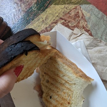 Underground Bakery - This is the prosciutto - A toasted sandwich with melted cheese and dark bread crust is held in a person's hand over a white paper tray. The sandwich appears to be partially eaten, revealing the melted cheese inside. The background features a colorful, patterned floor with geometric shapes and various colors, including red, green, and yellow. A paper napkin is also visible on the tray. - Bakeries Near Me - Dennis, Massachusetts