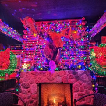 Camp Bar Third Ward - Santa Camp - A reindeer head with antlers is mounted on a wall decorated with multicolored Christmas lights. The fireplace below has a lit fire, and the surrounding area features holiday decorations, including wreaths and ornaments. The scene is festive and illuminated with vibrant, colorful lighting. - Bars Near Me - Milwaukee, Wisconsin