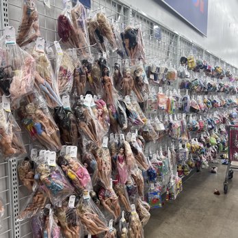 Unique - The image shows a retail store aisle with a wire grid wall filled with numerous dolls and toys in clear plastic bags. The dolls vary in size, style, and clothing, and are arranged in rows. To the right, shelves hold additional toys and accessories, with a shopping cart visible at the end of the aisle. The store has a bright, clean environment with a concrete floor and a large store sign partially visible at the top. - Thrift Stores Near Me - Silver Spring, Maryland