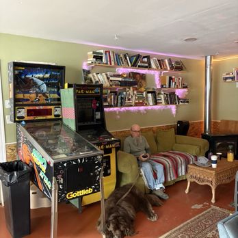 Nomad Cafe - A man with glasses sits on a green sofa holding a remote control, with a large dog lying on the floor in front of him. The room has bookshelves, a pinball machine, and a coffee table. - Coffee & Tea Near Me - Kanab, Utah