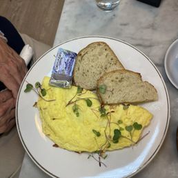 Omelette - A plate features a cooked omelette topped with microgreens, accompanied by two slices of bread. A small packet of butter is placed on the plate. The meal is set on a marble table with a glass of water and a napkin holder nearby. The setting appears to be in a cafe or restaurant.