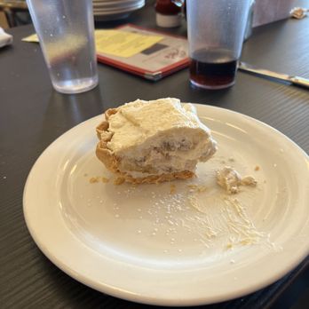 Joe's Dining - A slice of cream pie with a flaky crust and creamy filling is on a white plate, with some crumbs and filling remnants. Two glasses of water and a glass of dark soda are on the table. - Pizza Near Me - Santa Fe, New Mexico