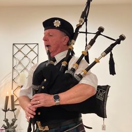 A man in Scottish dress, including a kilt and a hat, is playing the bagpipes in an indoor setting.