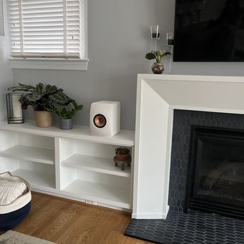 New Vision Construction - Living room corner with a white built-in shelf containing potted plants, a speaker, and decorative objects. A window with blinds is above the shelf. To the right, a fireplace with black hexagonal tiles surrounds the firebox. The room has hardwood flooring and neutral-colored walls. - Drywall Installation & Repair Near Me