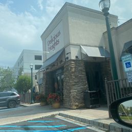 The image shows the storefront of Delicious Heights, featuring stone accents, potted plants, and outdoor seating, with parking spaces nearby.