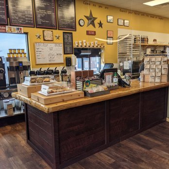 Cabin Coffee - The image shows the interior of a coffee shop with a wooden counter displaying baked goods in clear plastic covers. Behind the counter are coffee machines, a whiteboard with daily specials, and a yellow wall decorated with stars and framed items. The menu boards are mounted above the window, listing various drink options. The shop has a warm, inviting atmosphere with wooden flooring and a window providing natural light. - Coffee Roasteries Near Me - Ames, Iowa