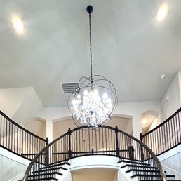 Light fixture - A chandelier with multiple lights hangs from a high ceiling in a large interior space featuring curved staircases and a balcony.