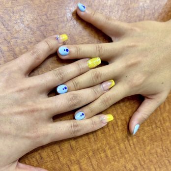 Master Nails - Laurel - Two hands with manicured nails are resting on a wooden surface. The nails are decorated with colorful designs, including blue, yellow, and white polish, with some nails featuring small embellishments and patterns. The image highlights the nail art and grooming, with the hands positioned to display the nails clearly. - Nail Salons Near Me - Laurel, Maryland