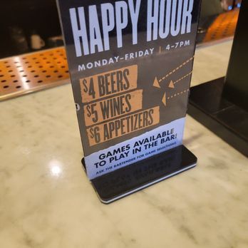 The Highbinder - Happy Hour prices - A small, freestanding tabletop sign promoting happy hour at a bar, with details about drink specials such as $4 beers, $5 wines, and $6 appetizers, available Monday through Friday from 4 to 7 PM. The sign also mentions that games are available to play in the bar, with a prompt to ask the bartender for game selections. The sign features bold text and a simple design, placed on a marble countertop. - Beer Bar Near Me - Washington, DC, District of Columbia