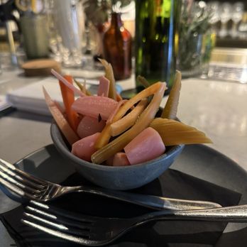 Root & Flower - Pickled veg - A small ceramic bowl contains pickled vegetables such as radishes, carrots, and possibly celery or other roots, arranged on a black napkin. The table setting includes two forks placed parallel to each other. In the background, there are bottles, glassware, and decorative elements, indicating a restaurant or bar environment with ambient lighting. - Wine Bars Near Me - Vail, Colorado