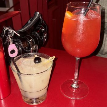 R Bar - Saturn Punch (left) and Rialto Spritz (right) - A creamy cocktail with a cherry garnish and a large red drink with ice are on a red table in a bar or restaurant. - Cocktail Bars Near Me - Tucson, Arizona