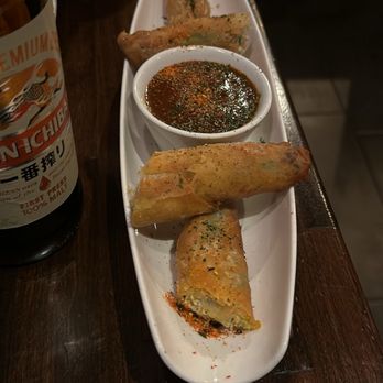 Koze Teppan Grill - Avocado Eggrolls - Four spring rolls with a dipping sauce are arranged on a white oval plate on a dark wooden table in a restaurant setting. - Sushi Bars Near Me - El Paso, Texas