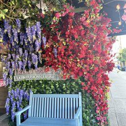 A blue wooden bench is positioned against a wall decorated with purple and red flowering plants outside a Latin fusion cuisine restaurant.
