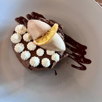3128 Social House - Chocolate dessert - A dessert featuring a chocolate base topped with dollops of whipped cream, a lemon slice, and two scoops of ice cream. The plate has a drizzle of chocolate sauce on the side, and the dessert is presented on a plain white plate, with a wooden surface visible in the background. - Gastropubs Near Me - Albuquerque, New Mexico